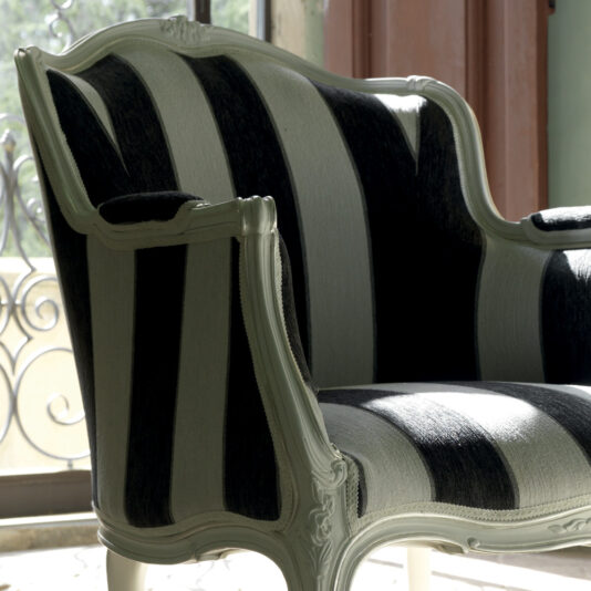 The Venetian Style White Italian Armchair features an ornate wooden frame and is upholstered in black and white striped fabric. This vintage chair is placed near a window with ironwork details, allowing natural light to illuminate its elegant curves and texture.