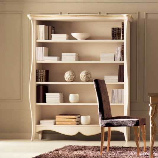 A large beige wooden bookcase filled with various books, decorative items, and a couple of vases stands against a light-colored wall. In front of the Large Italian Luxury Freestanding Bookcase is a plush, brown upholstered chair on a beige carpet, near a wooden table edge.