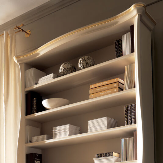 A Large Italian Luxury Freestanding Bookcase, featuring elegant gold trim and a tall, beige design, is filled with neatly organized books and decorative items. White and black ceramic spheres adorn the top shelf, while white blank CD cases are interspersed among the books. Cream curtains hang beside the bookcase.