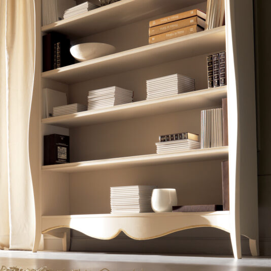 The Large Italian Luxury Freestanding Bookcase is filled with neatly arranged stacks of books and a few decorative items. A white ceramic bowl graces the second shelf, while a small white mug sits on the bottom shelf. Sunlight streams in from the left, illuminating the elegant setup.