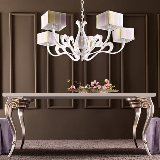 A modern dining room features the Designer Italian Dining Table Set, showcasing a white rectangular table adorned with ornate silver legs and a decorative floral centerpiece. Above the table hangs a white chandelier with five cube-shaped lampshades, all set against a dark brown paneled wall.