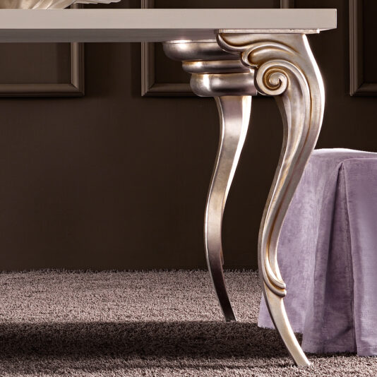 Designer Italian Dining Table Set A close-up of an elegant, ornate table leg with a metallic finish and intricate curves in a classical style. The Designer Italian Dining Table Set is set on a gray carpet next to a mauve upholstered piece, perhaps a chair or an ottoman. The background features a dark-colored wall.