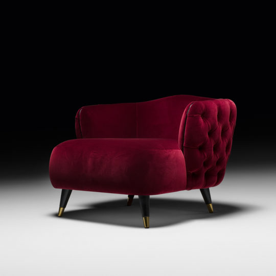 A luxurious Button Upholstered Designer Velvet Tub Chair, featuring a deep red velvet finish with a tufted back and sides, stands against a dark background. The chair's black wooden legs adorned with golden tips add an elegant and sophisticated touch.