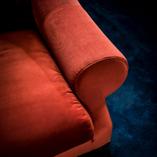 Close-up of the armrest and cushion of the Classic Designer Velvet Armchair against a dark blue carpeted floor. The rich textures contrast beautifully, highlighting the luxurious fabric of the chair and the deep color of the carpet.