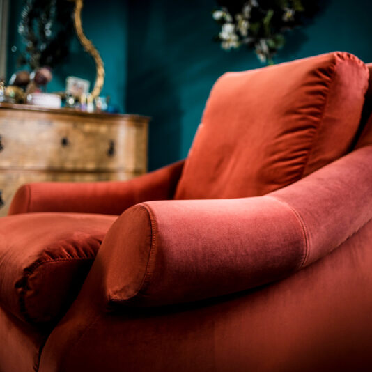 A close-up shot of the Classic Designer Velvet Armchair features its plush, red fabric in a cozy, stylish living room. In the blurred background, a wooden dresser and an ornate mirror with gold accents add a vintage touch to the scene.