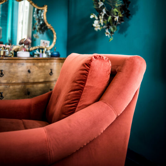 A cozy, plush Classic Designer Velvet Armchair sits against a teal wall in an elegantly decorated room. A vintage wooden dresser with various items and a large, ornate gold-framed mirror are in the background, along with a leafy plant arrangement hanging on the wall.