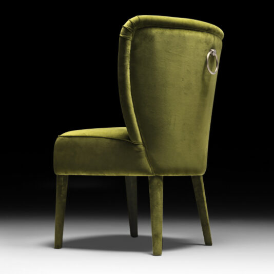 Designer Velvet Dining Chair The Designer Velvet Dining Chair, featuring a green velvet upholstery and a high backrest, stands against a black background. Its dark wooden legs and sleek, modern design are complemented by an elegant silver ring detail on the back.