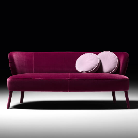 The High End Velvet Day Sofa, upholstered in plush magenta velvet with tapered legs, includes two light lavender circular pillows positioned on the right side, all set against a black background.