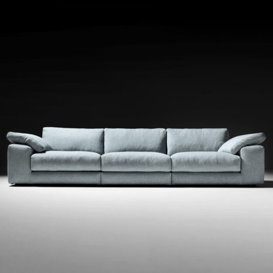 The Large Linen Designer Modular Sofa, boasting a modern light blue hue with three seat cushions and padded armrests, is showcased against a dark, gradient background. This designer sofa features a low-profile design and exudes both comfort and spaciousness.