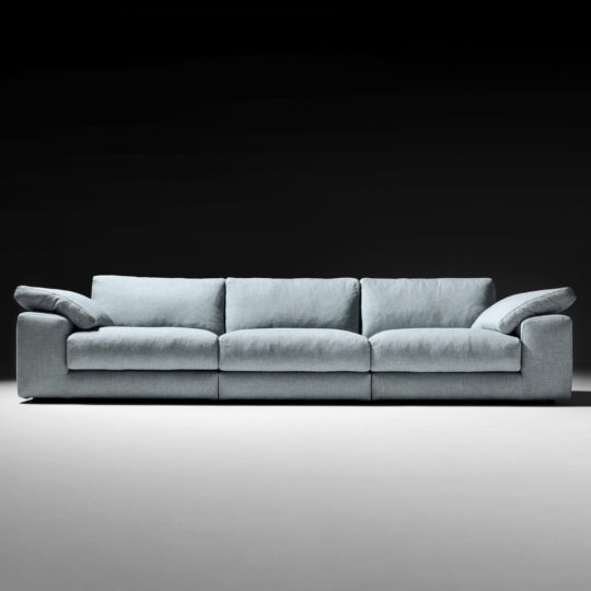 The Large Linen Designer Modular Sofa, boasting a modern light blue hue with three seat cushions and padded armrests, is showcased against a dark, gradient background. This designer sofa features a low-profile design and exudes both comfort and spaciousness.
