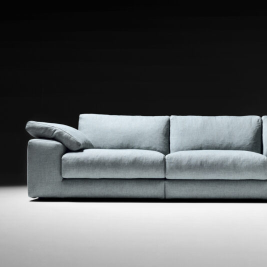 The Large Linen Designer Modular Sofa, featuring a modern and minimalistic design, is set against a black background. It boasts light-gray upholstery and large plush cushions, with one back cushion and a seat cushion partially visible on the far right. This versatile piece of contemporary furniture highlights its sleek linen material.