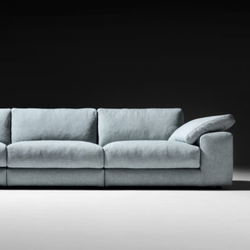 Large Linen Designer Modular Sofa The Large Linen Designer Modular Sofa, featuring a modern and minimalist design, showcases plush light gray cushions against a black background. This spacious three-seater sofa includes one side with a gently reclined backrest, sitting on a light-colored floor to create a striking contrast.