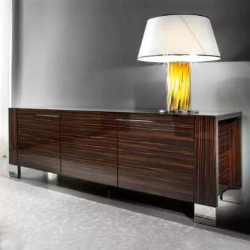 Luxury Italian Wenge Buffet A stylish Luxury Italian Wenge Buffet with a sleek, glossy finish stands against a gray wall. It features three compartments and is supported by metallic legs. On top of the buffet, there is a chic lamp with a white shade and a yellow, textured base. The floor is covered with a light-colored carpet.