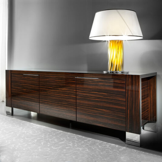 A stylish Luxury Italian Wenge Buffet with a sleek, glossy finish stands against a gray wall. It features three compartments and is supported by metallic legs. On top of the buffet, there is a chic lamp with a white shade and a yellow, textured base. The floor is covered with a light-colored carpet.