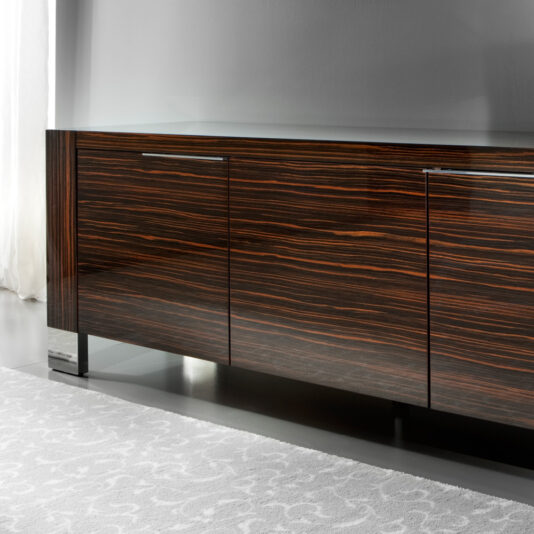 Luxury Italian Wenge Buffet A sleek, modern Luxury Italian Wenge Buffet with a dark wood veneer and subtle grain patterns features two metal handles on its doors. It stands on thin, black legs in a minimalist, light-colored room, where a white curtain is partially visible.