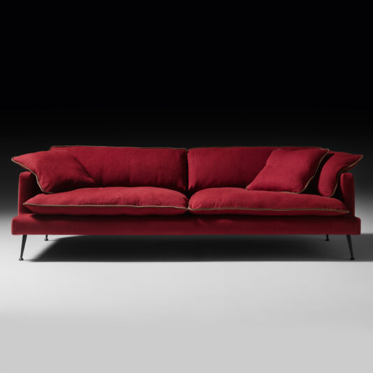 The Modern Linen Designer Sofa, upholstered in a deep red linen, stands out against a black background with its plush cushions and elegant thin gold piping detail. The sofa's sleek, straight lines and slim black metal legs lend it a contemporary and stylish look.