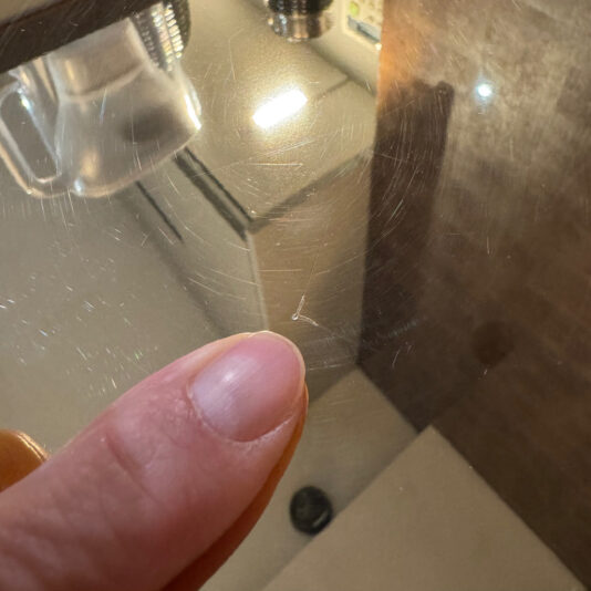 A finger points to a small scratch on reflective glass, with kitchen cupboards, a tap, and the sleek silhouette of the Contemporary Gold Designer Side Table mirrored in the surface.