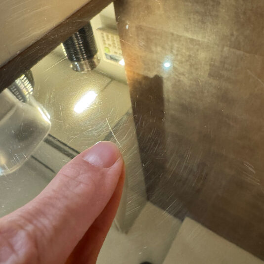 A close-up shows a hand pointing at a thin scratch on the shiny surface of the Contemporary Gold Designer Side Table, with overhead lights and nearby objects clearly reflected in its lustrous finish.