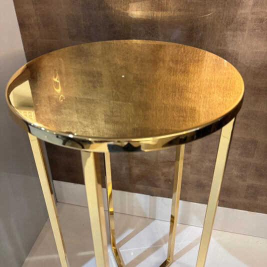 The Contemporary Gold Designer Side Table features a small, round, reflective tabletop and slender legs. It stands on a white floor against a brown textured wall, adding modern elegance to any space.