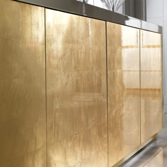 Close-up of the Large High End Gold Leaf Sideboard featuring shiny metallic gold doors and a glossy dark countertop, reflecting light in a contemporary interior.