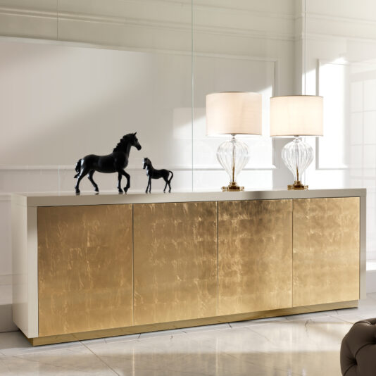 A Large High End Gold Leaf Sideboard with four doors displays two glass table lamps with white shades and two black horse figurines in a modern, bright room featuring marble floors and reflective walls.