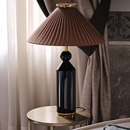 The Classic Designer Luxury Table Lamp, featuring a pleated brown lampshade and a sleek black base, is elegantly placed on a round metallic side table. Positioned next to a window adorned with beige curtains and set against an intricately patterned wall, it enhances the room's decor with its sophisticated charm.