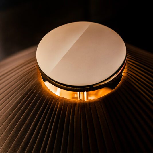 Close-up of the Classic Designer Luxury Table Lamp featuring a large, circular, metallic disc at the top. The lamp emits a warm, orange light through a grooved, dark brown shade below the disc, creating an ambient, softly lit scene.