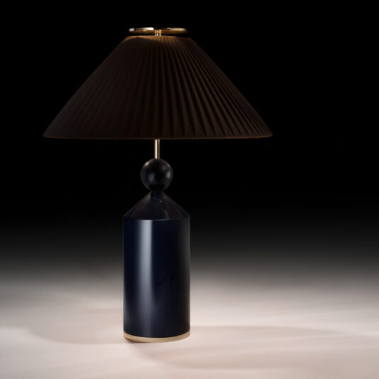 The Classic Designer Luxury Table Lamp features a dark cylindrical base adorned with gold accents and is topped with a pleated black lampshade. Its minimalist design casts a muted, elegant light against a dark background, exuding modern sophistication.