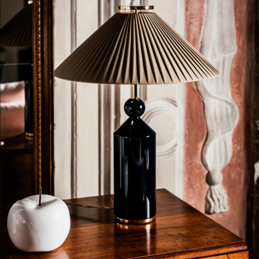 A black Classic Designer Luxury Table Lamp with a beige pleated lampshade sits on a wooden table next to a white decorative apple-shaped object. The background features an ornate wall with a mirrored section reflecting part of the scene.