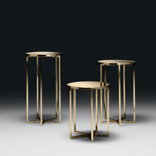 A set of three Contemporary Gold Designer Side Tables features sleek, geometric metal frames and round tabletops. The tables are arranged in a slightly staggered formation against a dark background, accentuating their reflective surfaces and minimalist design.