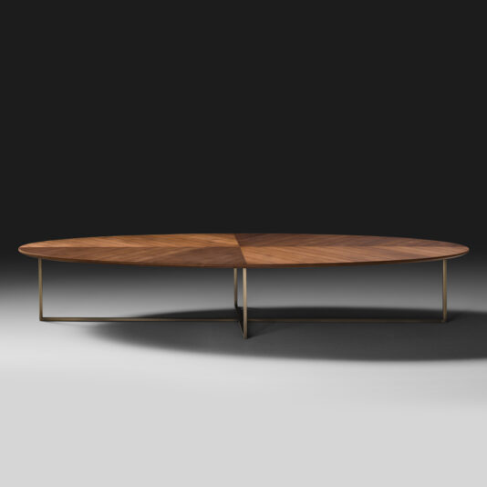 The Contemporary Walnut Oval Coffee Table has a wooden top with angled, triangular inlays and is supported by thin, minimalist metal legs. Set against a dark background, the sleek and modern design stands out beautifully.