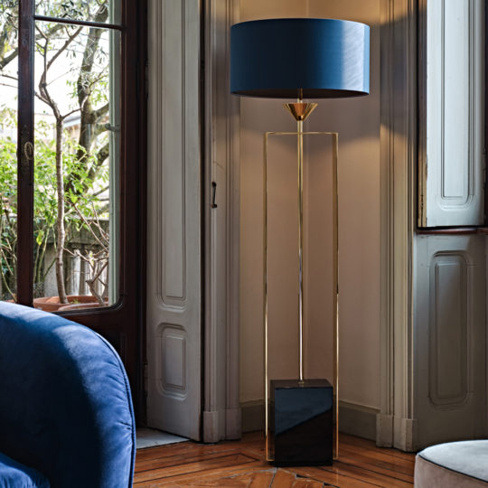 A stylish living room corner is highlighted by the Gold Contemporary Designer Floor Lamp, which features a modern blue lampshade, a sleek gold stand, and an elegant black marble base. The room is adorned with wooden flooring, and in the foreground, a plush blue velvet sofa adds to the luxurious ambiance. Large windows with wooden shutters complete this sophisticated setting.