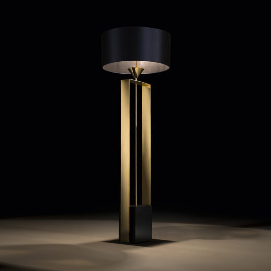 Gold Contemporary Designer Floor Lamp The Gold Contemporary Designer Floor Lamp features a sophisticated, modern design with a tall, rectangular gold base and an oversized, cylindrical black lampshade that emits a soft, warm light. This exquisite piece stands against a dark background, casting a subtle glow on the floor.