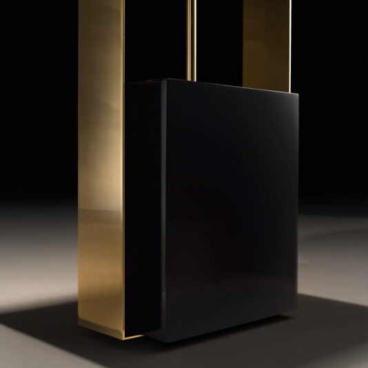Gold Contemporary Designer Floor Lamp The Gold Contemporary Designer Floor Lamp showcases a large, rectangular black block elegantly suspended between two tall, vertical golden columns against a dark background. The sleek surfaces reflect light, offering a minimalist and stylish appearance.