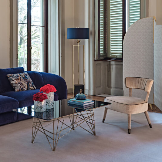 Gold Contemporary Designer Floor Lamp A modern living room featuring a blue velvet sofa, a beige accent chair, and a glass-top coffee table with a geometric metal base. The table holds vibrant red flowers and books. In the background are tall windows with shutters, the Gold Contemporary Designer Floor Lamp, and a partition screen.