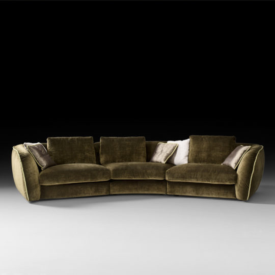 The Curved Designer Velvet Modular Sofa, featuring a plush olive-green velvet finish with thick cushions and armrests, is centered against a black background. Accent pillows in various shades of cream and brown are arranged on the sofa. The minimalist setting highlights the curved furniture’s luxurious texture and design.