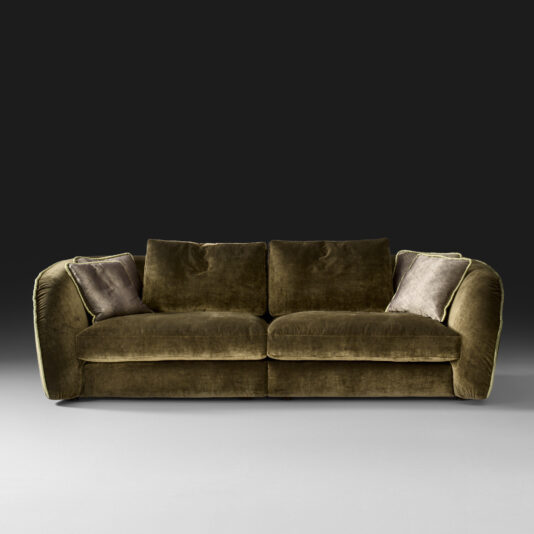A plush, Designer Velvet Modular Sofa in dark olive-green, featuring two large seat cushions and back cushions, is centered against a dark gray background. The sofa has rounded armrests and is adorned with two light beige throw pillows, one on each side.