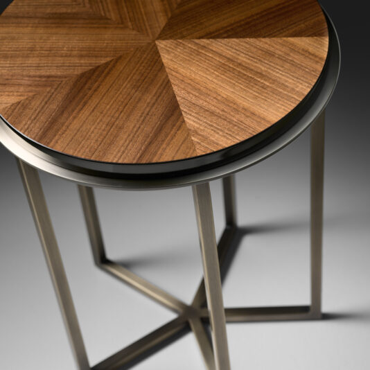 High End Walnut Veneered Side Table The High End Walnut Veneered Side Table showcases a small, round top adorned with an intricate geometric wood veneer pattern, resting gracefully on slender steel legs that epitomize minimalist design. The table's surface radiates a rich, natural walnut finish, while the legs offer a sleek metallic look.