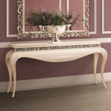The Luxury Cream And Silver Console Table, featuring intricate carvings and gracefully curved legs, is set against a mauve wall. Adorned with a decorative silver floral arrangement on its surface, an expansive ornate mirror mounted above reflects the beauty of the arrangement.