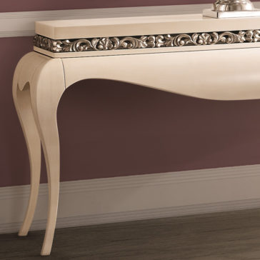 This Luxury Cream and Silver Console Table boasts ornate, intricate carvings on its upper front edge. With elegantly curved legs, it stands against a mauve wall with white trim and baseboards. The table's surface is minimalist, adorned with a small square decorative item.
