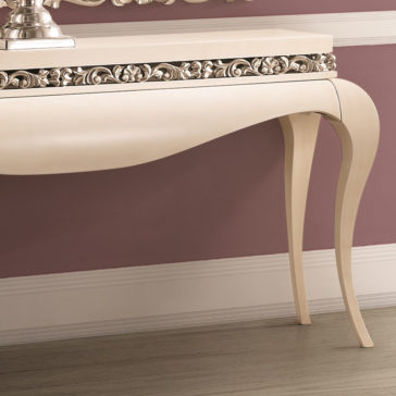 The Luxury Cream And Silver Console Table, featuring curved legs and intricate floral carvings, is positioned in front of a dusty rose-colored wall with white trim. Boasting a stylish, vintage design, the table adds an elegant touch to the room.