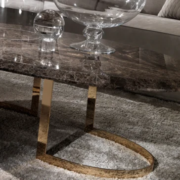 Modern 24 Carat Gold Plated Oval Marble Coffee Table In a lavish living room, a Modern 24 Carat Gold Plated Oval Marble Coffee Table graced with a glass orb and bowl stands out. Its shimmering gold metallic legs shine on a plush gray carpet, complemented by an elegant gray couch.