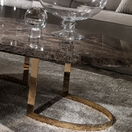 In a lavish living room, a Modern 24 Carat Gold Plated Oval Marble Coffee Table graced with a glass orb and bowl stands out. Its shimmering gold metallic legs shine on a plush gray carpet, complemented by an elegant gray couch.