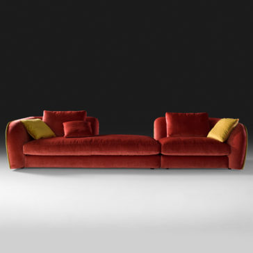 A sleek, red Chaise Style Velvet Designer Modular Sofa with plush, rounded cushions set against a black backdrop. The designer sofa includes two chaise-style lounges on either end and offers an array of coordinated cushions, featuring one yellow and one red pillow on each side. The floor underneath is pristine in white.