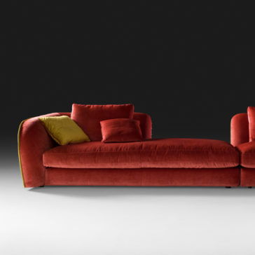A modern Chaise Style Velvet Designer Modular Sofa in rich red velvet with plush cushions against a dark backdrop. The sofa features three cushions in vibrant hues: one red, one yellow, and one orange, neatly arranged. Designed with a left armrest and sleek contemporary lines, it epitomizes modular sofa elegance.