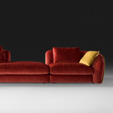 A stylish, contemporary red Chaise Style Velvet Designer Modular Sofa set against a dark background. This L-shaped piece features smooth lines and plump cushions, with one contrasting yellow cushion adding a pop of color. The setting is modern and minimalist.