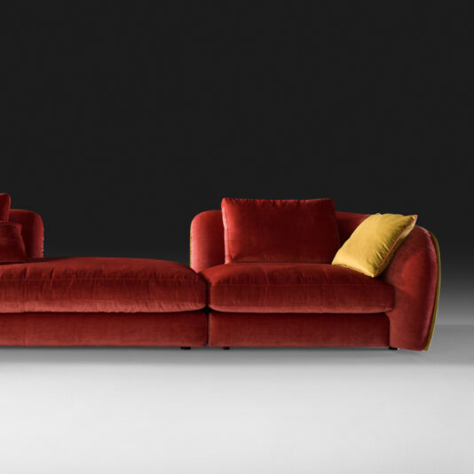 Chaise Style Velvet Designer Modular Sofa A stylish, contemporary red Chaise Style Velvet Designer Modular Sofa set against a dark background. This L-shaped piece features smooth lines and plump cushions, with one contrasting yellow cushion adding a pop of color. The setting is modern and minimalist.