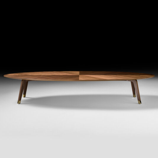 The Large Designer Walnut Oval Coffee Table features angled legs with brass caps and is set against a dark background. The tabletop showcases a geometric wood grain pattern, elevating its sophistication.