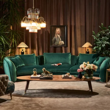 A luxurious living room boasts an emerald green velvet sofa, a large chandelier, and a Large Designer Walnut Oval Coffee Table adorned with decorative items like a vase of flowers. A portrait on the wall is framed by plants and elegant lamps.