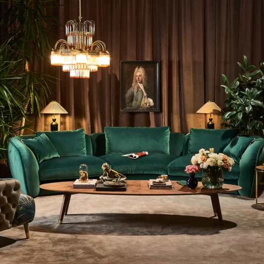 Large Designer Walnut Oval Coffee Table A luxurious living room boasts an emerald green velvet sofa, a large chandelier, and a Large Designer Walnut Oval Coffee Table adorned with decorative items like a vase of flowers. A portrait on the wall is framed by plants and elegant lamps.